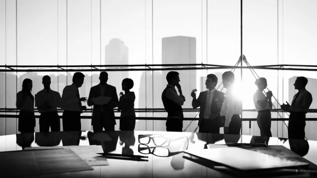 Silhouettes of people talking in a glass-walled meeting room, with glasses and papers on the table in front
