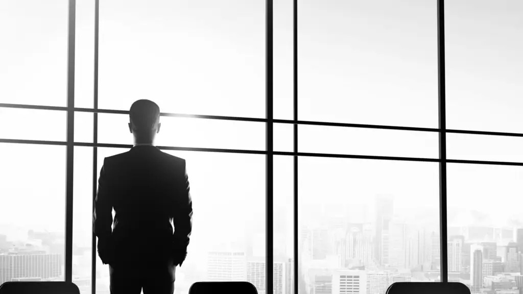 Black-and-white photo of a man in a suit standing at a boardroom window, looking out over a city